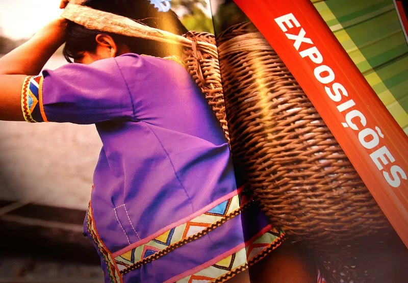 Detalhe da revista do Encontro Niterói América do Sul na seção Exposições: mulher de blusa roxa bordada com cesto de palha.